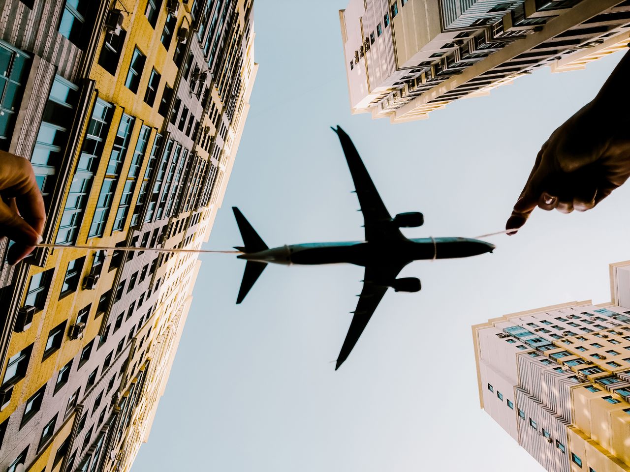 A toy airplane held with string appears flying between high-rise buildings against the sky.