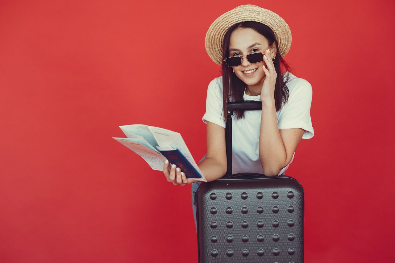 Delighted young woman in straw hat in casual clothes leaning on suitcase and checking tickets before traveling on red background