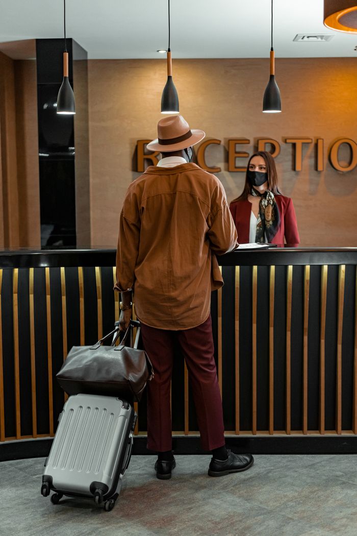 Home : SRL Tours & Travel - Travel The World With Our Eyes Man with luggage checking in at hotel reception. Staff wearing mask, indoors, travel theme.